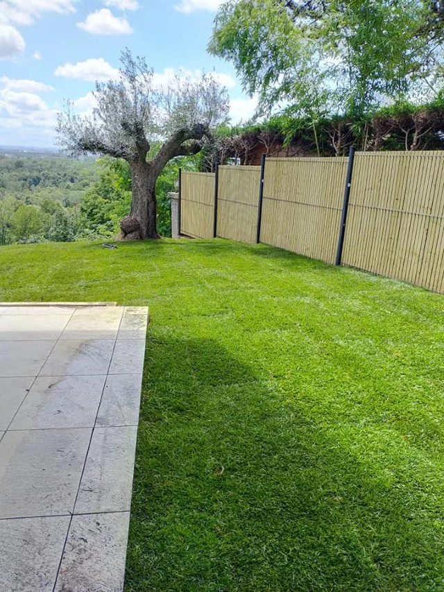 Espace extérieur couvert avec salon de jardin en bois exotique disposé en L, coussins noirs, suspensions en rotin tressé au plafond